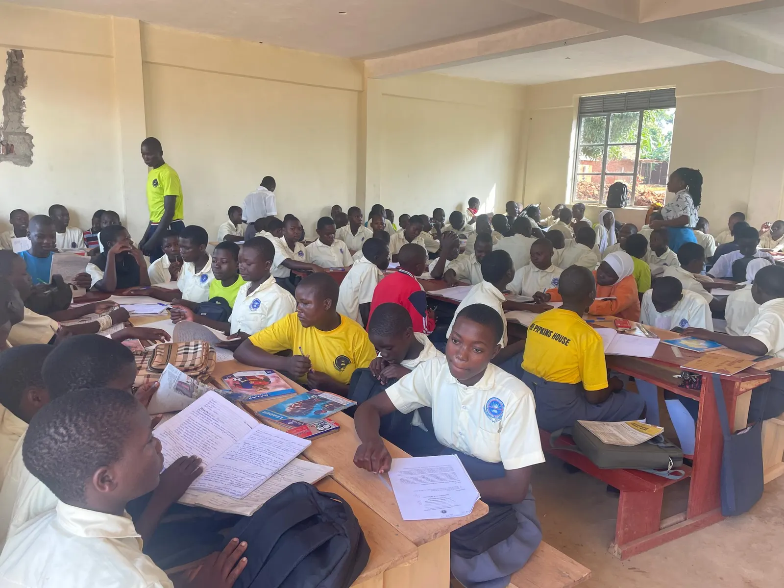 Students read exam papers at wooden desks while a teacher stands nearby