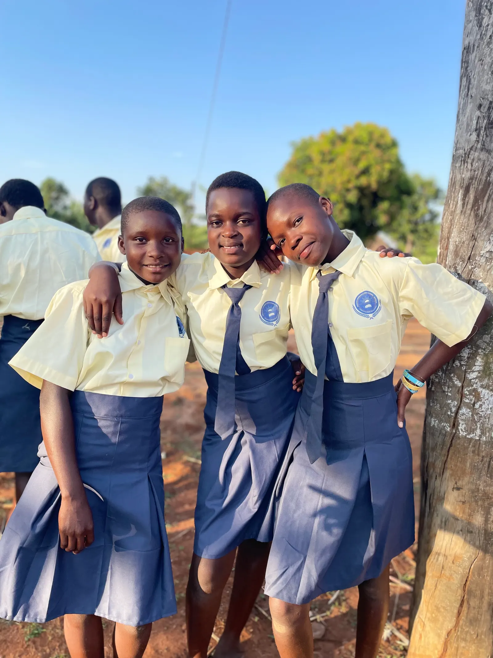 Three students in yellow shirts and navy skirts standing arm in arm beside a tree