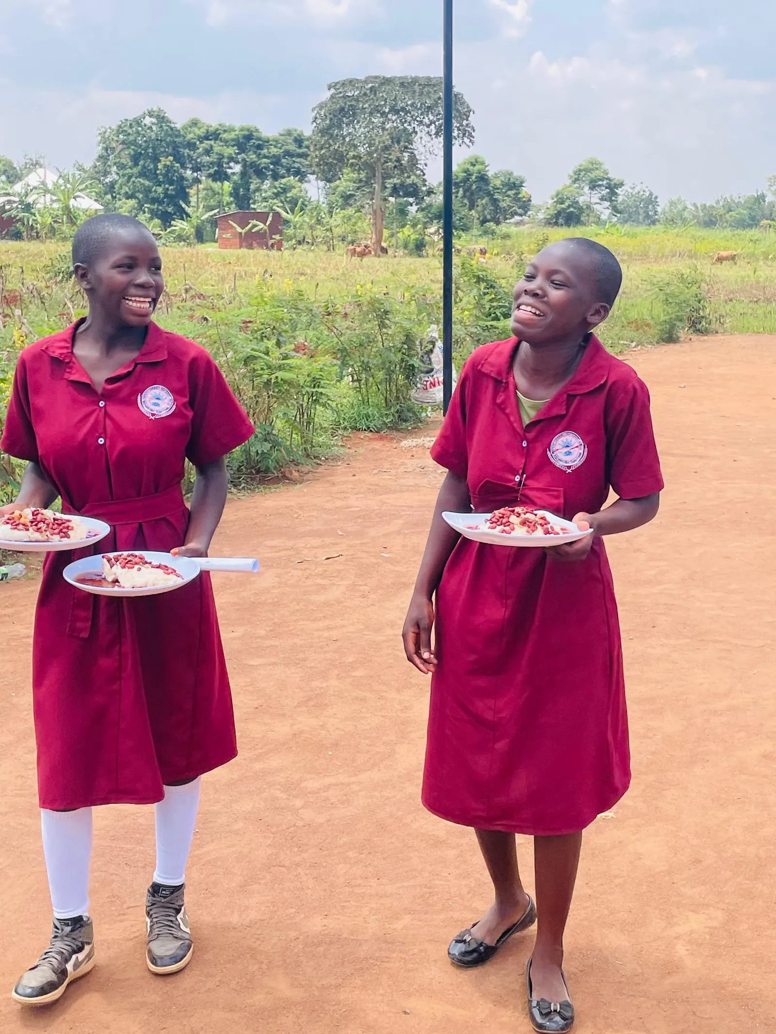 Two students carry plates of food across the school compound