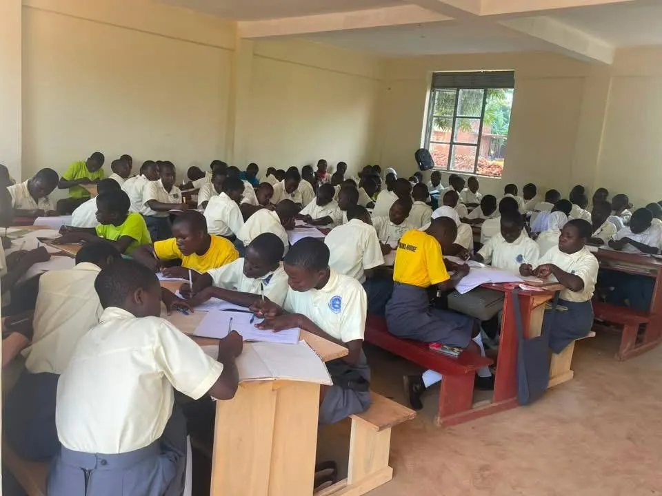 Students in yellow and white shirts sit in rows at wooden desks in a long hall