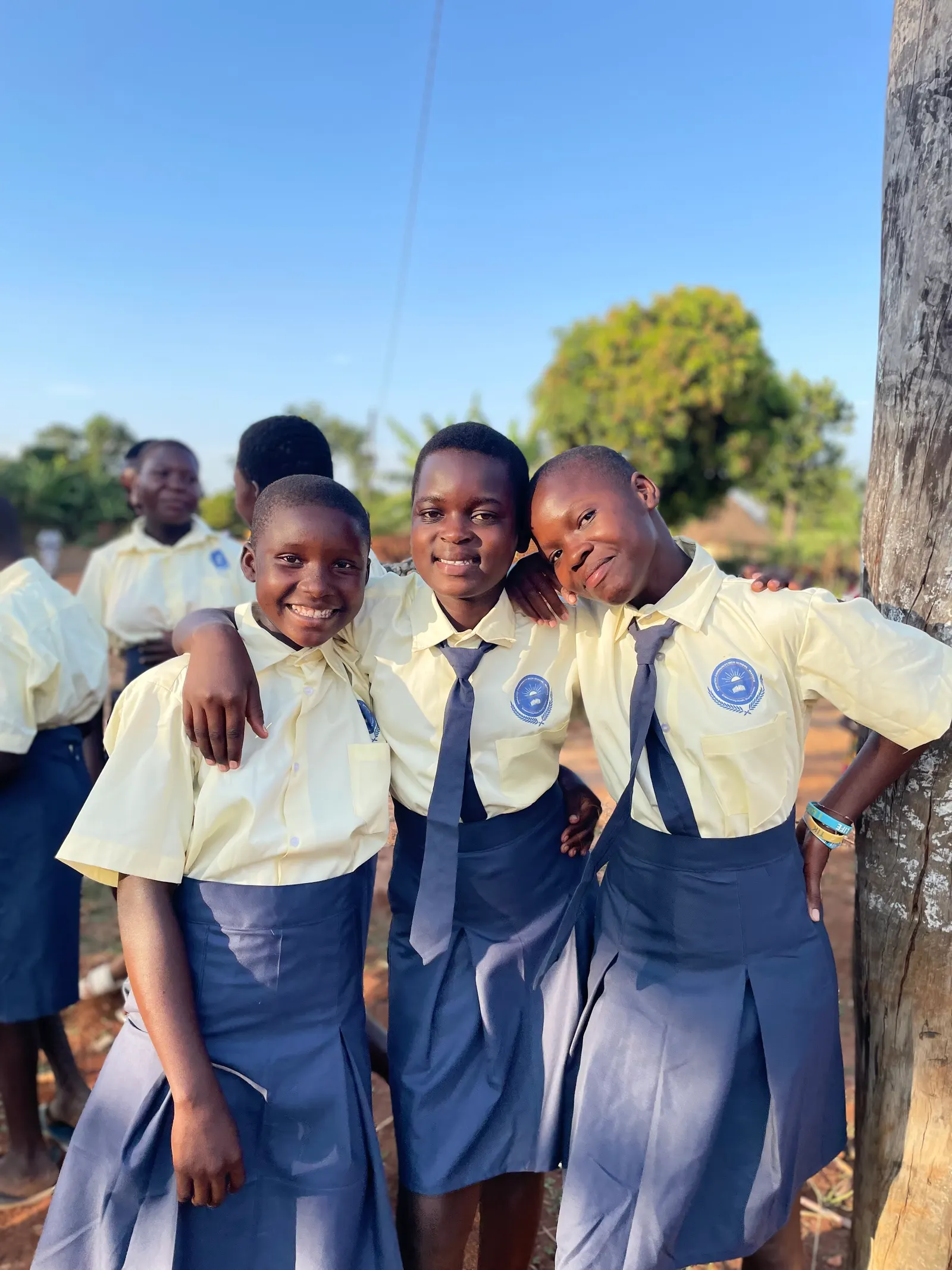 Three girls in uniform lean together and smile beside a wooden pole